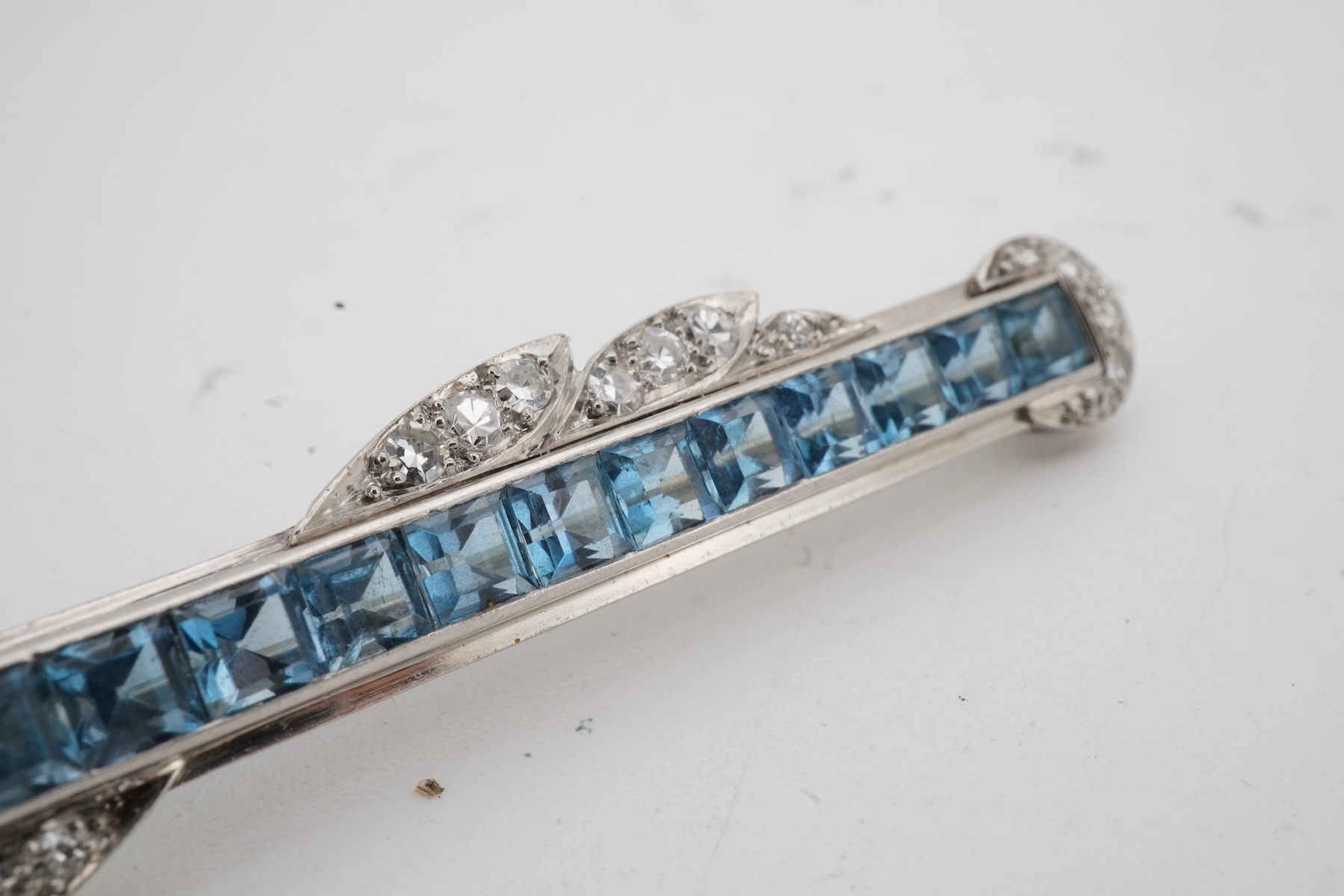 An Art Deco Aquamarine and diamond brooch, 1930s, of bar design, set with calibré-cut aquamarines, accented with foliate scrolls set with single-cut diamonds, length 5.5cm, white metal unmarked, gross weight 8 grams